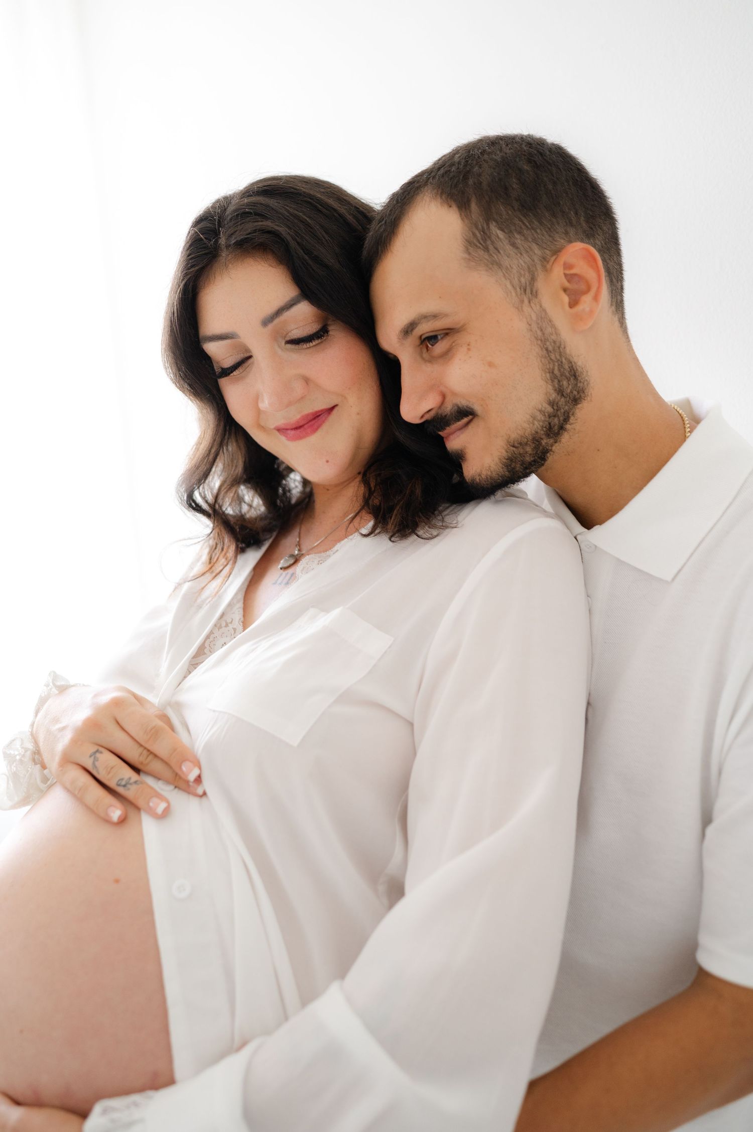Intimate maternity photos showing a couple in white shirts against a bright background.