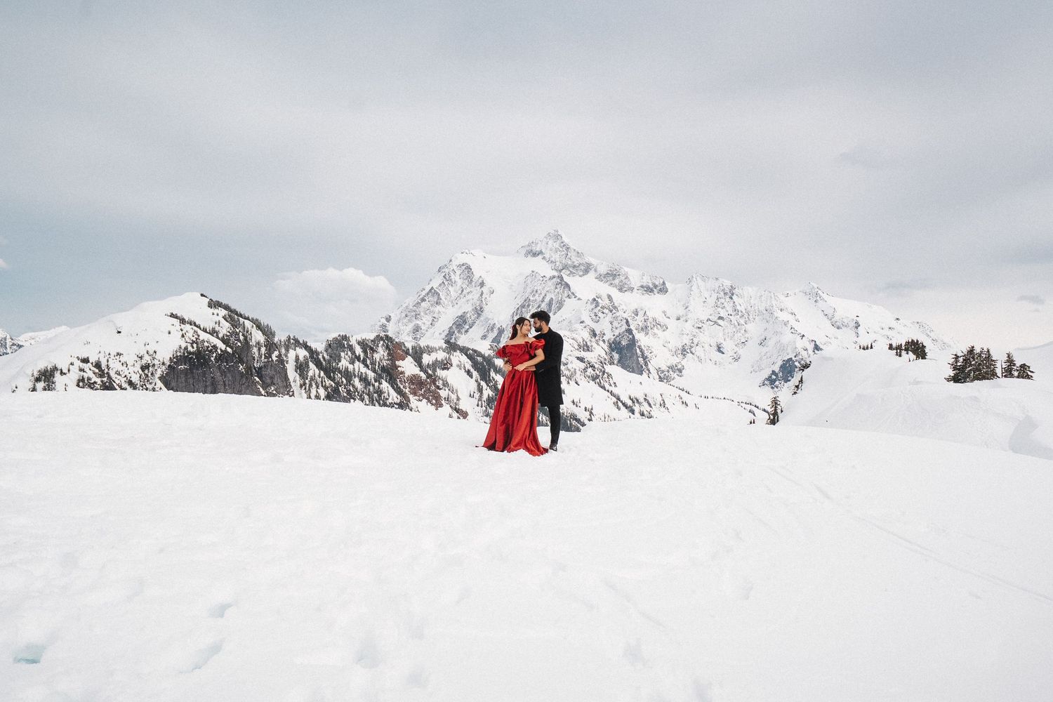 Two figures in red stand on a snowy mountain peak with dramatic snow-capped mountain ranges in the background.