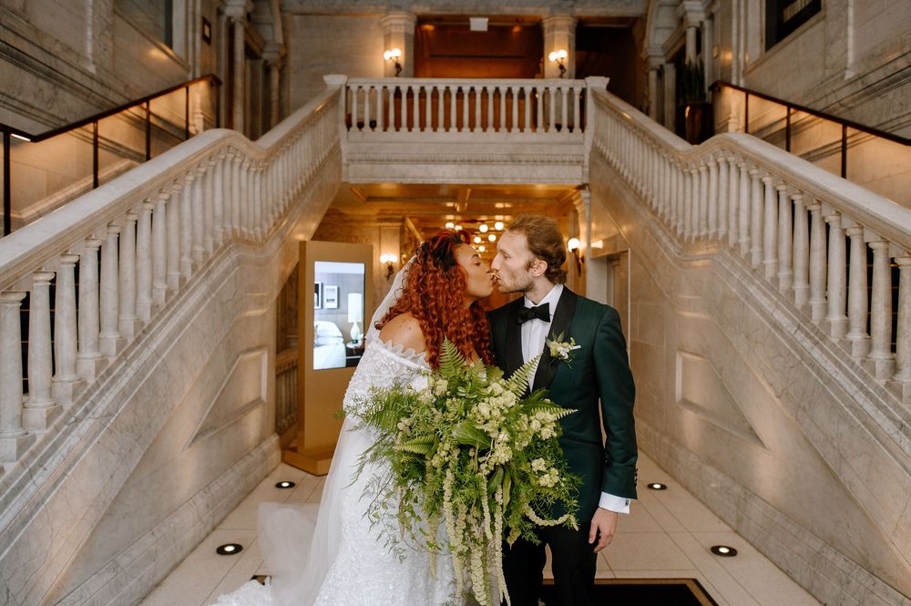 Two wedding attendees share an intimate moment on an elegant marble staircase with classical white bannisters.