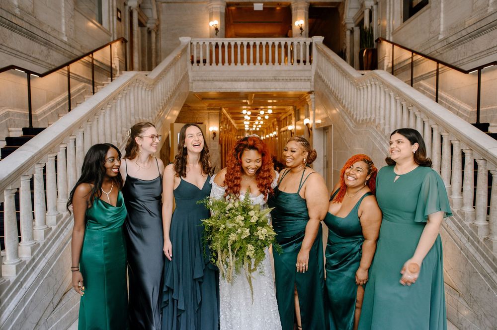 Wedding party wearing dark green and navy dresses share candid joyful moments in a grand staircase setting.