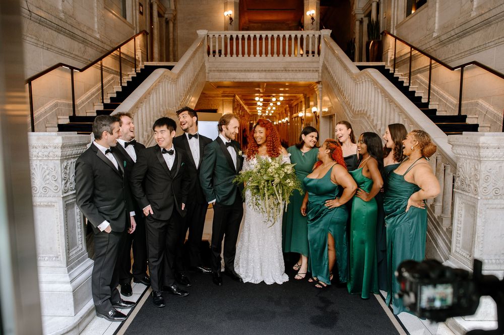 Wedding party in elegant black tuxedos and emerald green dresses pose together on grand staircase at venue.