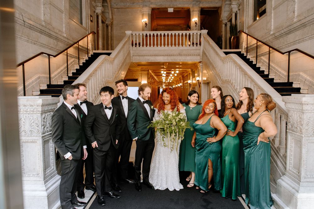 A wedding party stands in an elegant white hallway flanked by grand pillars and staircase during a formal celebration.