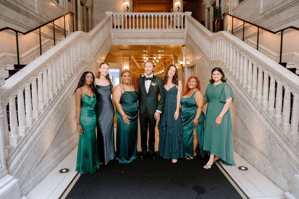 A wedding party poses in green formal dresses and black tuxedo on an elegant white marble staircase.