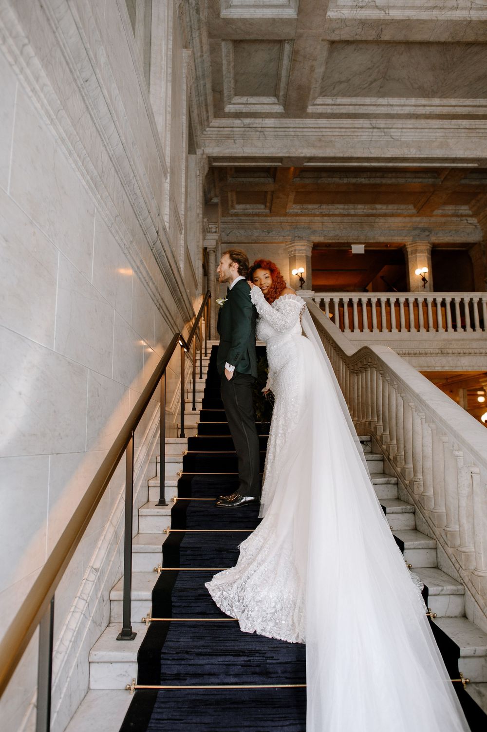 A bride in a flowing white gown with a long train walks up an elegant marble staircase with white railings.