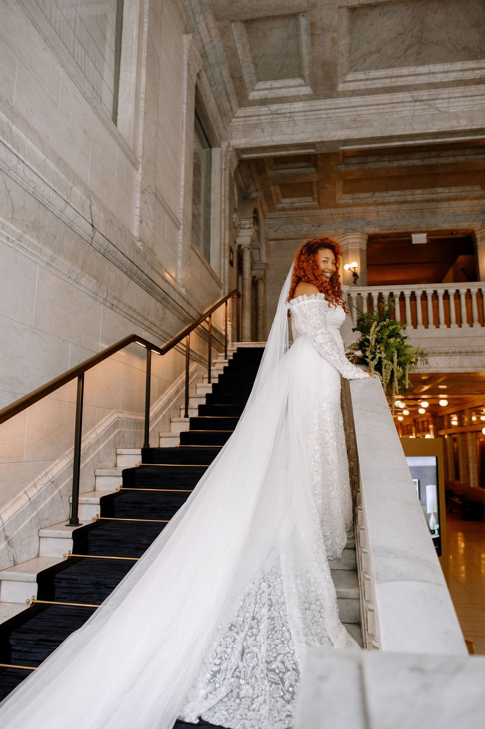 A flowing lace wedding gown with long train displayed on an elegant marble staircase with black carpet runner.