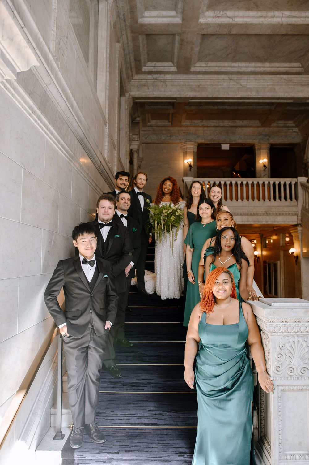 Wedding party members in formal attire and green dresses walking up a grand marble staircase.