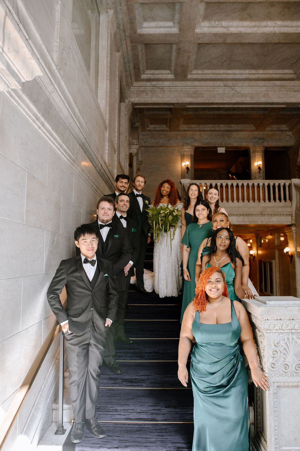 Wedding party members in elegant attire pose on a grand staircase in a historic building.