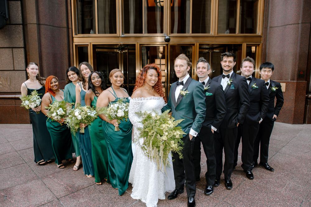 Wedding party in emerald green dresses and black tuxedos pose together outside stone building entrance.
