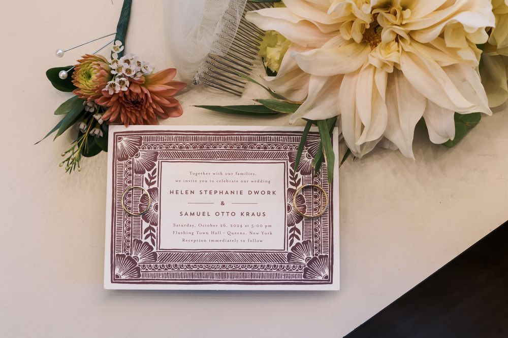 Elegant wedding invitation with ornate border design displayed next to a pale peach dahlia flower.