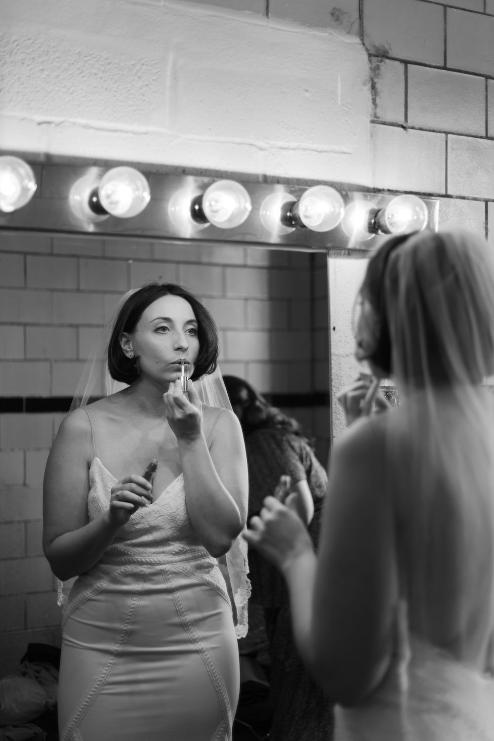 A person in a white dress applies lipstick in a mirror with vanity lights.