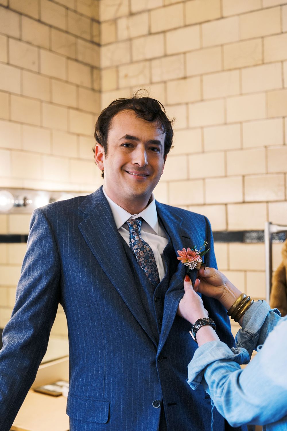 Someone adjusts a boutonniere on a navy blue suit jacket against a cream brick wall background.