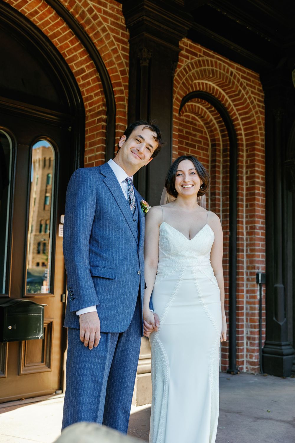 A formal wedding portrait outside a brick building with arched architecture.