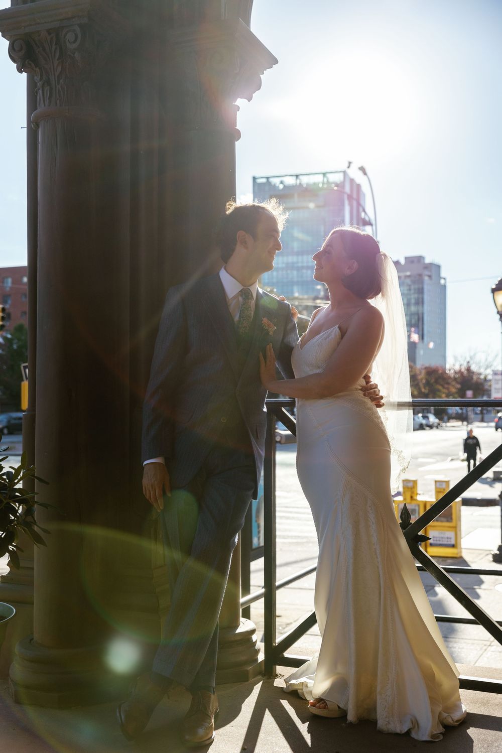 A couple in wedding attire poses against a sunlit column outdoors in an urban setting.