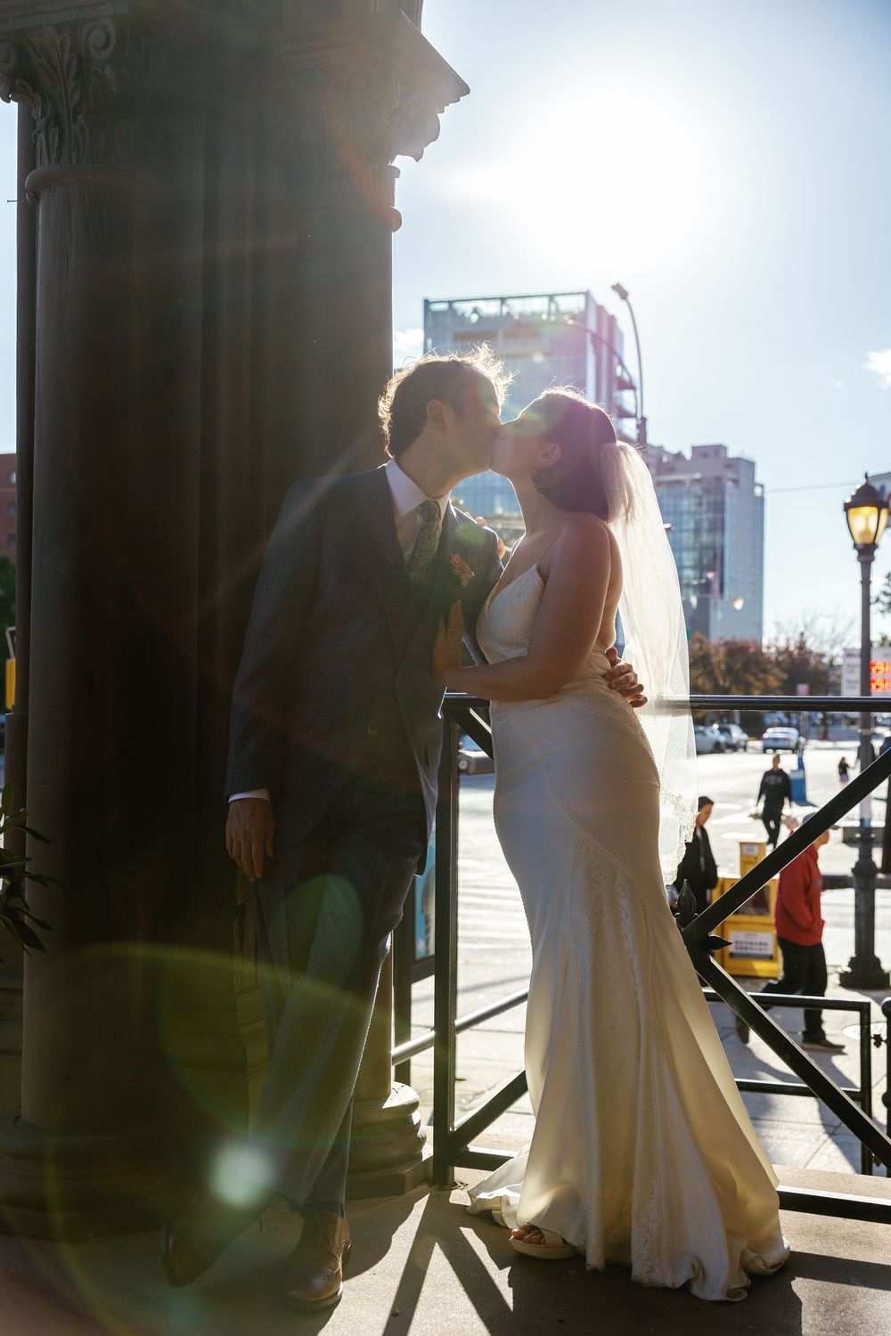 A romantic wedding couple shares a kiss on a sunlit balcony with iron railings and an ornate column.