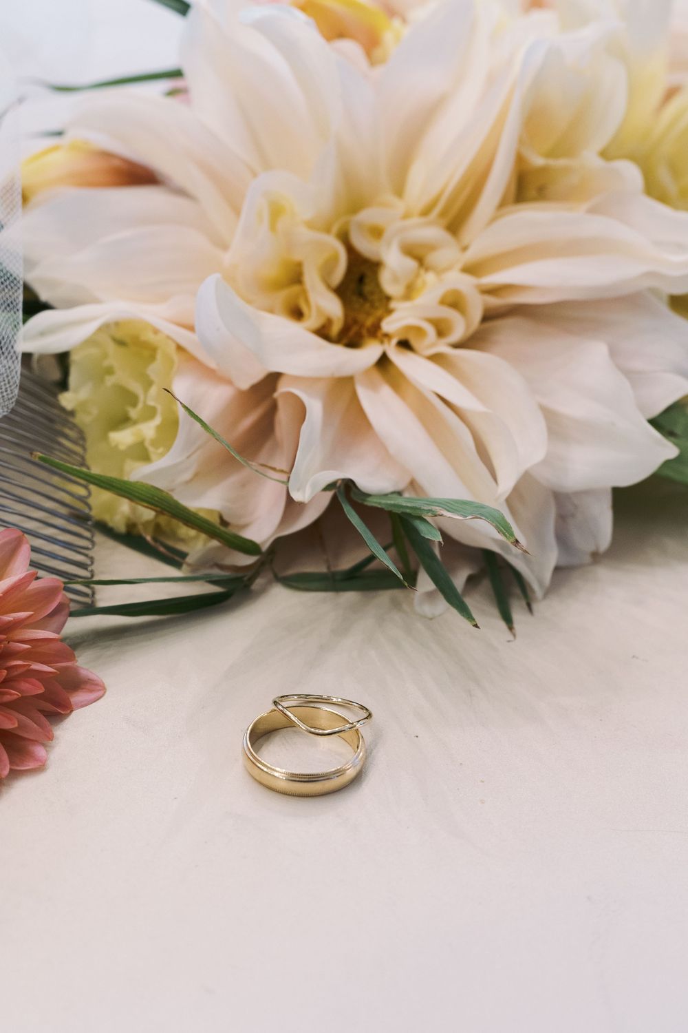 Wedding rings displayed next to soft pink dahlia flowers on white surface.