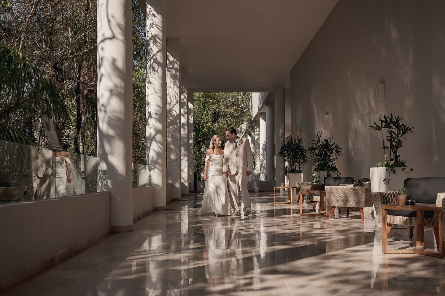 Sun casts dramatic shadows through pillars onto a bright corridor with rattan furniture and potted plants.