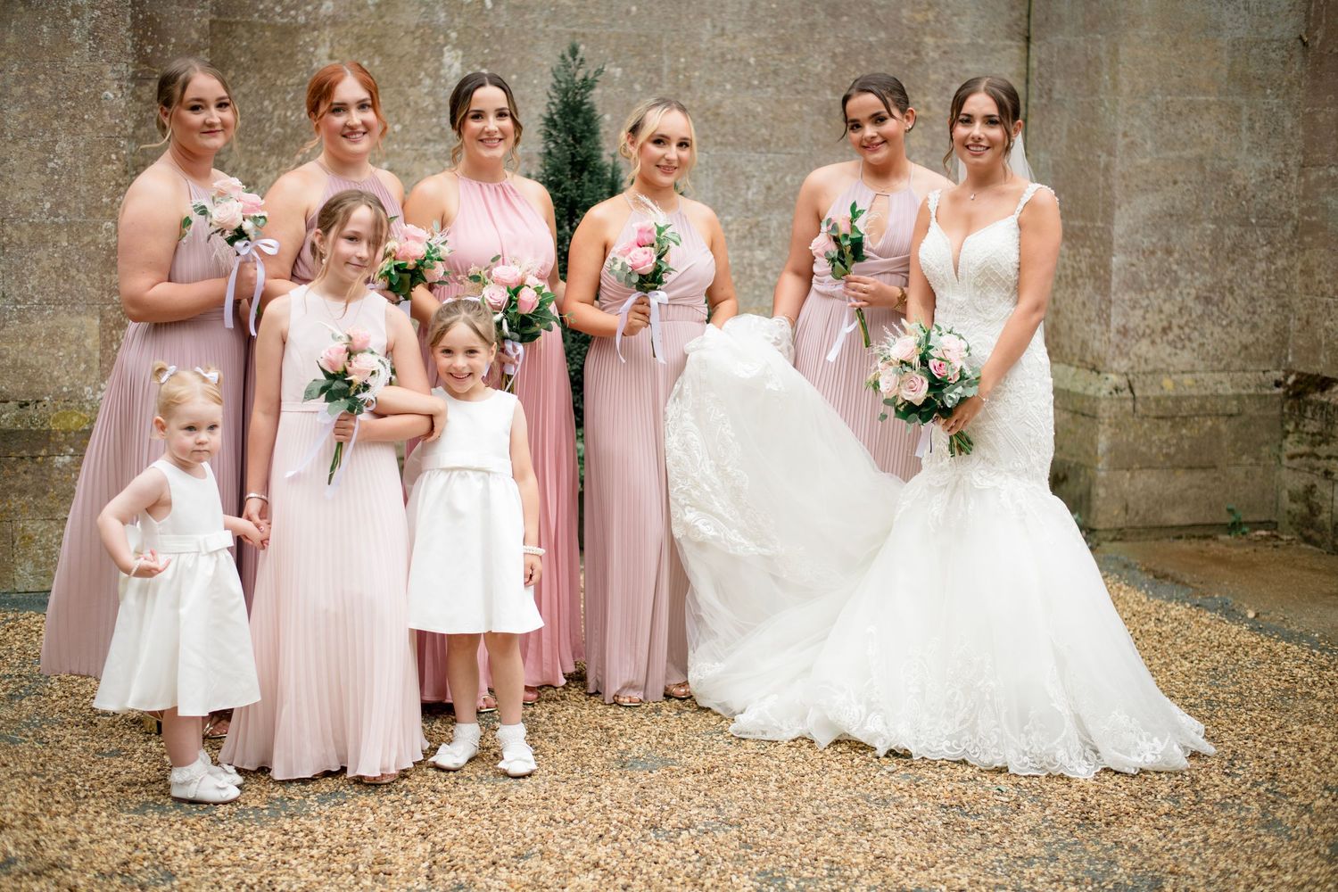 A wedding party poses in pink bridesmaids dresses and white gown against a stone wall at an elegant outdoor ceremony.