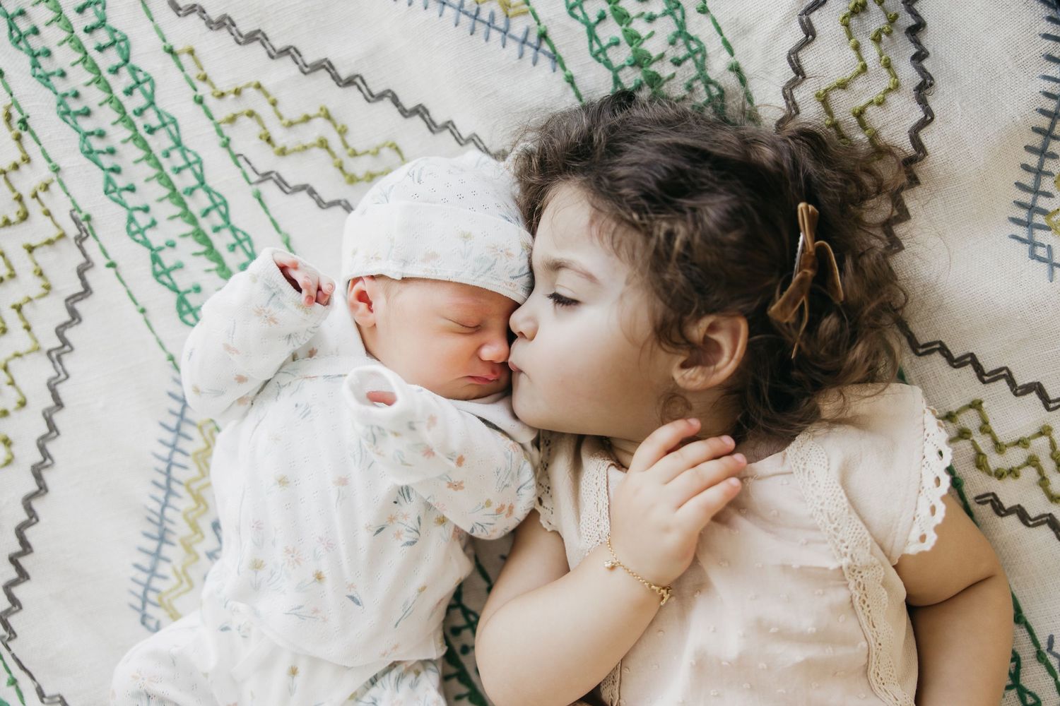 Two children lying together on a patterned blanket with one giving the other a sweet kiss.