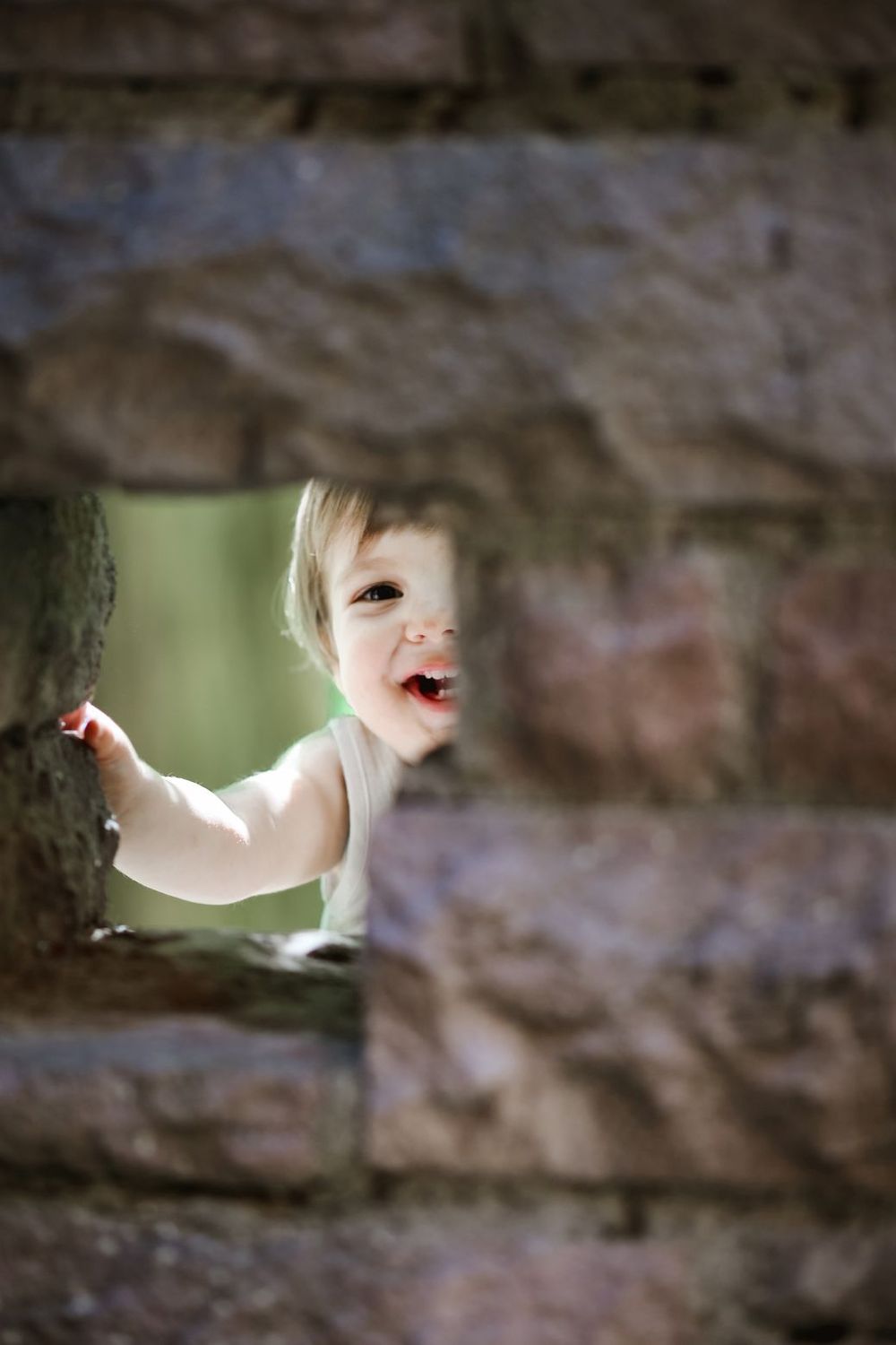 A playful child peeks through a hole in a stone wall with a bright smile.