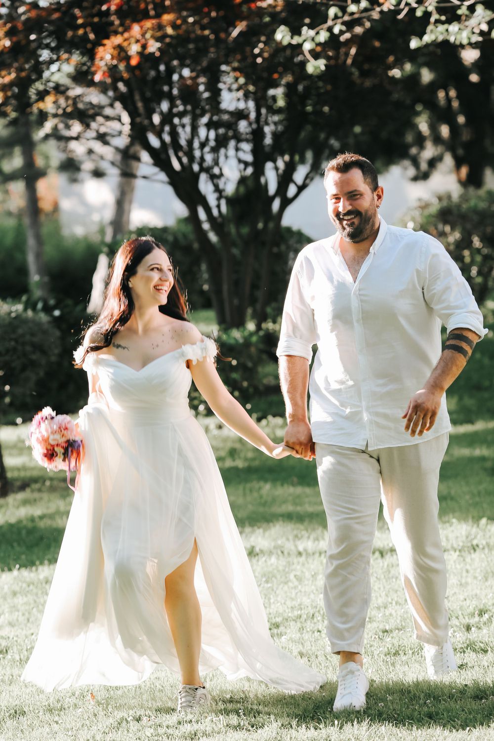 A couple in white attire holds hands while walking on grass during their outdoor wedding photoshoot.