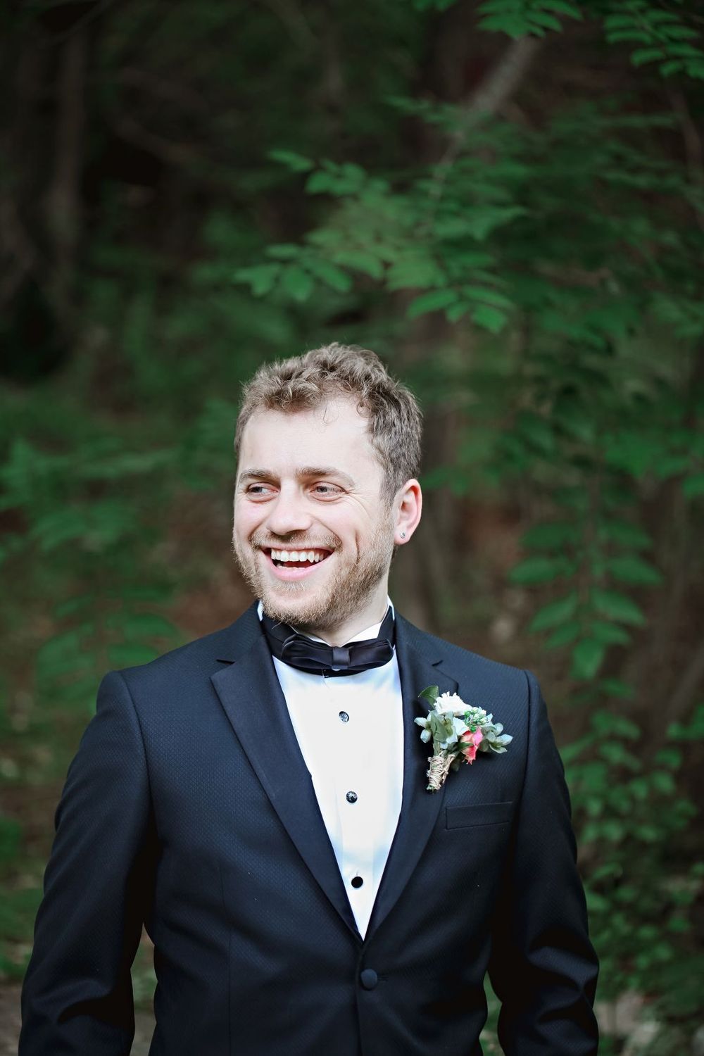 A groom in a black tuxedo with boutonniere smiles against a blurred forest backdrop.
