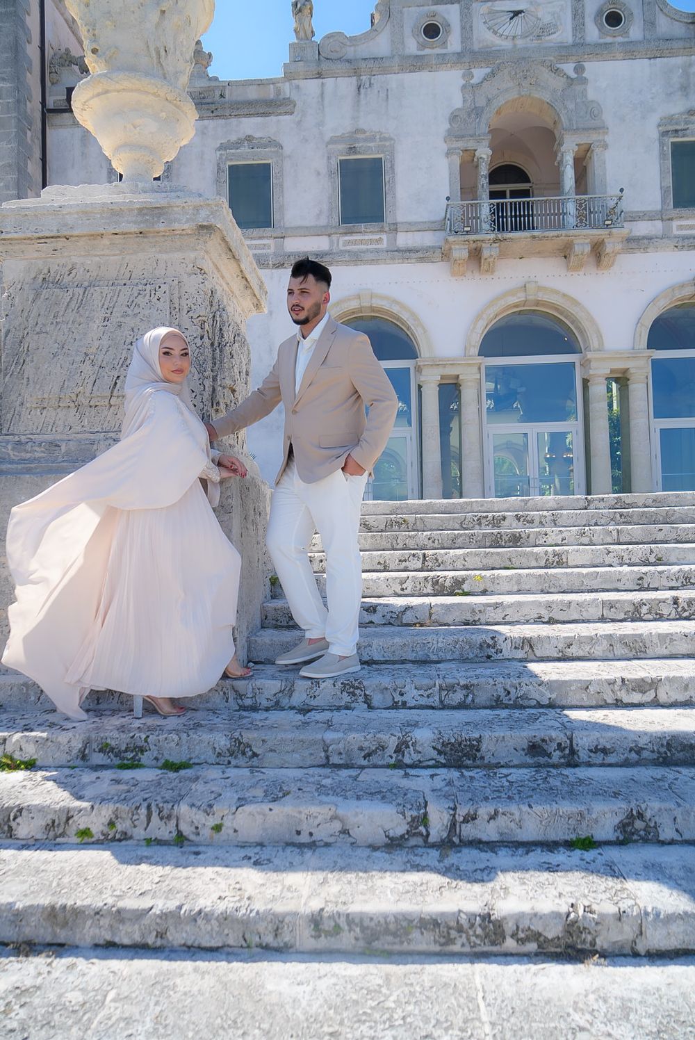 A couple in elegant white attire poses on marble steps of a historic building, woman in hijab holding man's hand.