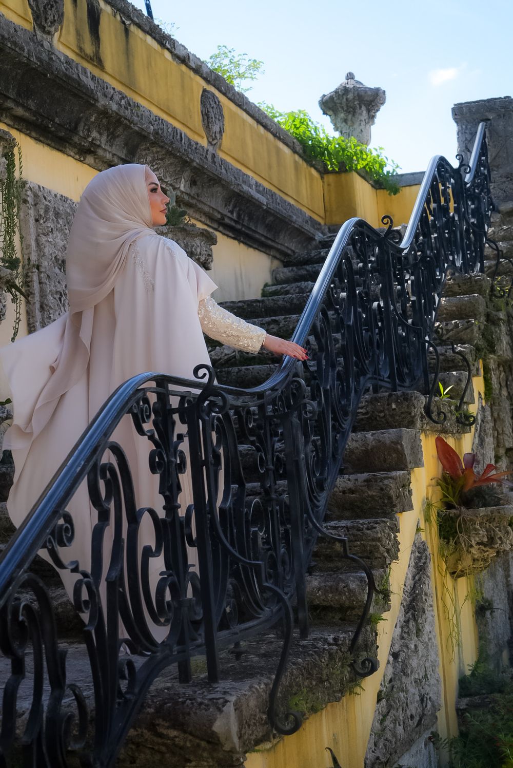 Woman in beige hijab and abaya ascending ornate iron staircase at historic stone building.