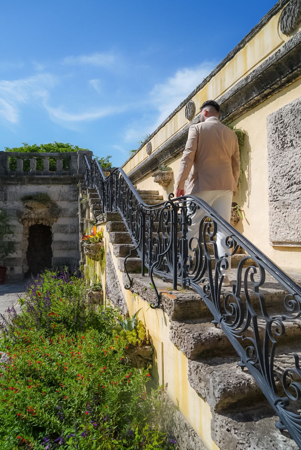 Man in beige jacket ascending ornate iron-railed stone stairs at a historic Mediterranean villa with lush gardens.