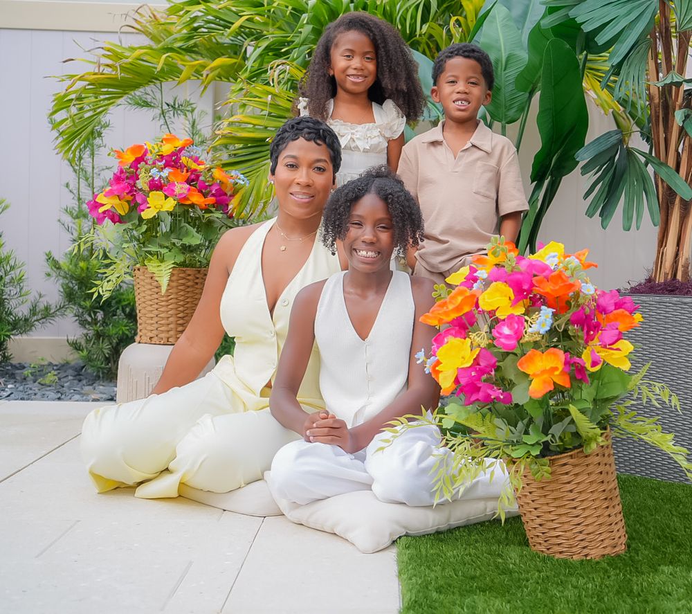 A woman and three children pose together outdoors surrounded by vibrant tropical floral arrangements in wicker baskets.