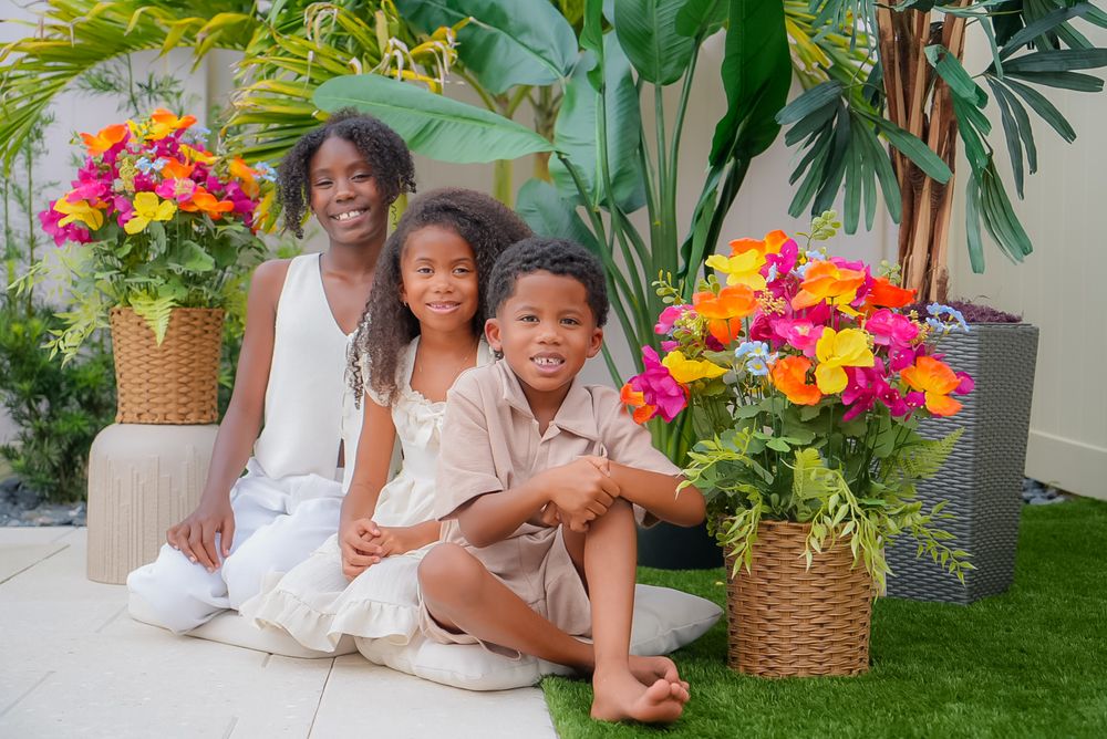 A woman and two children sit together smiling outdoors, surrounded by vibrant tropical flowers and lush green plants.