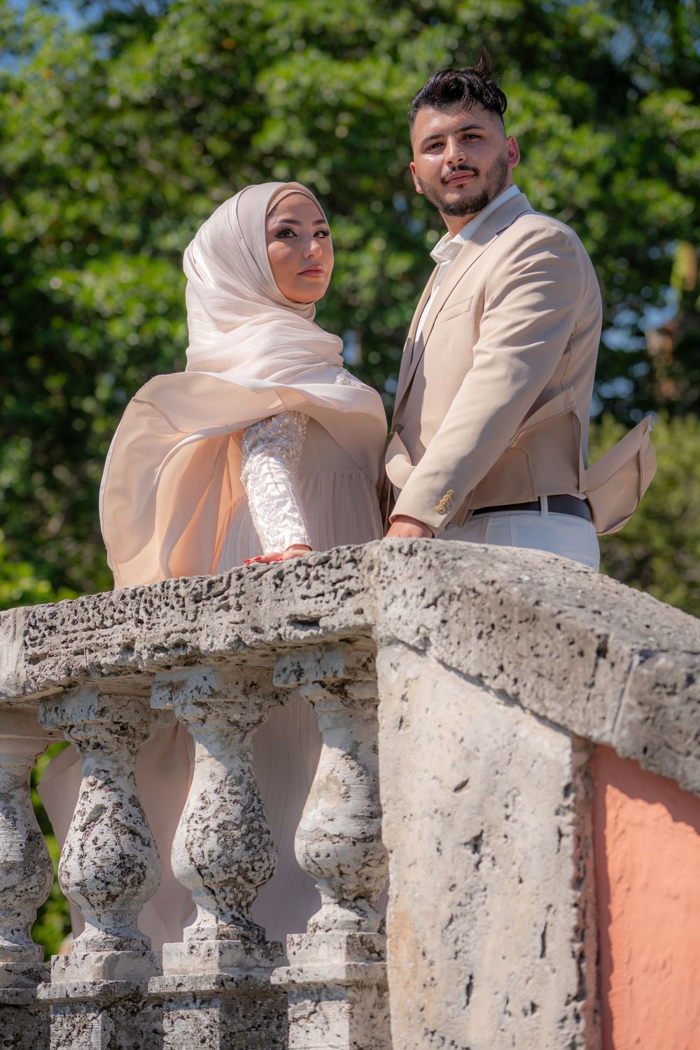 A couple in elegant beige attire poses on a stone balcony surrounded by lush greenery, the woman wearing a hijab.