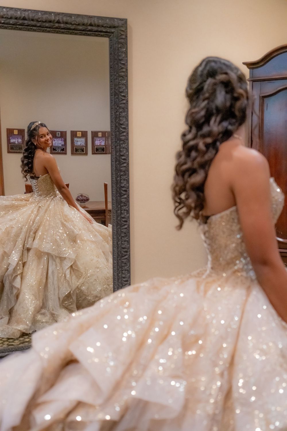 A bride in a sparkling champagne ball gown admires her reflection in an ornate floor mirror, showcasing her elegant curled updo.