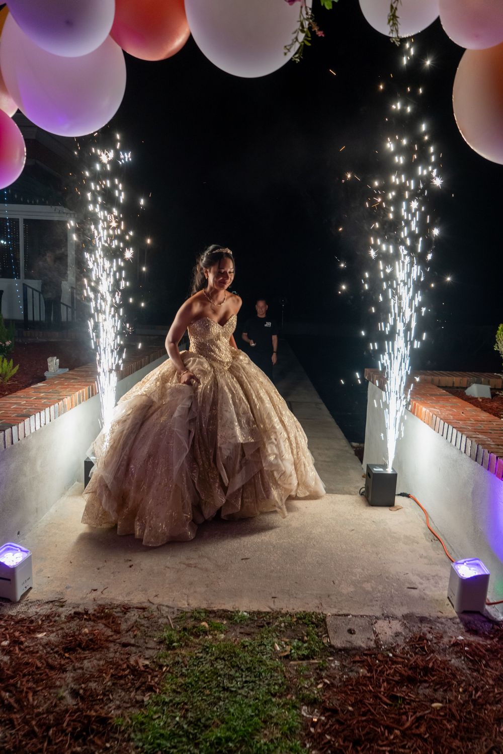 Young woman in a gold ball gown walks between cold spark fountains under colorful balloons at a quinceañera celebration.