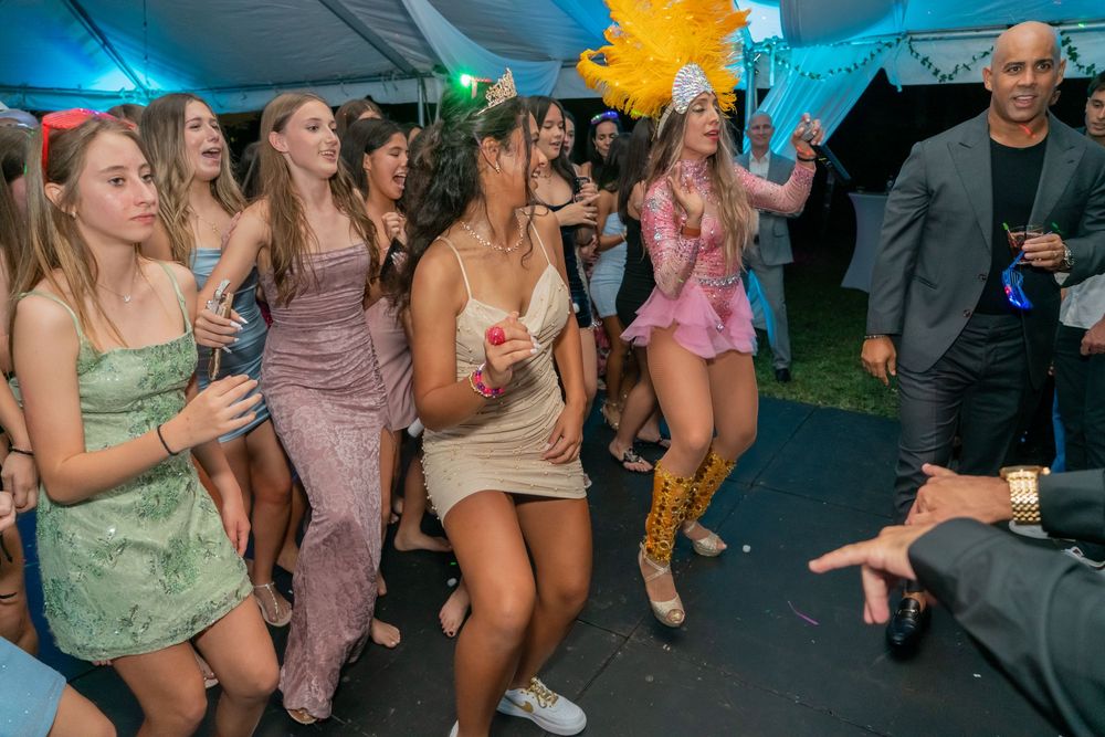 Young women in elegant dresses dancing at a festive outdoor party under a tent with colorful decorations.