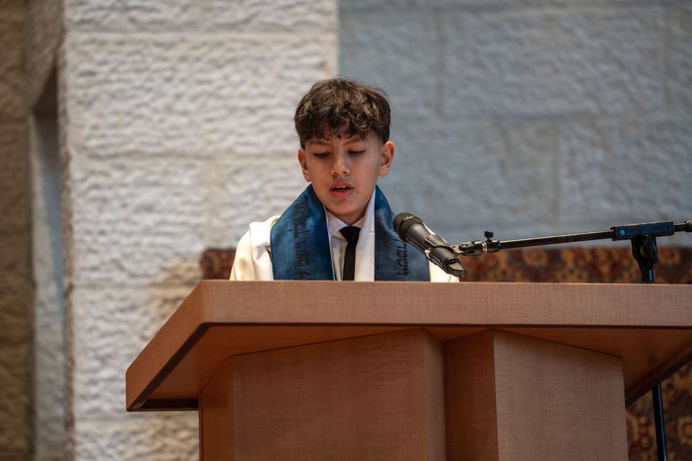 Young boy reading at a wooden podium wearing a blue tallit during a Bar Mitzvah ceremony.