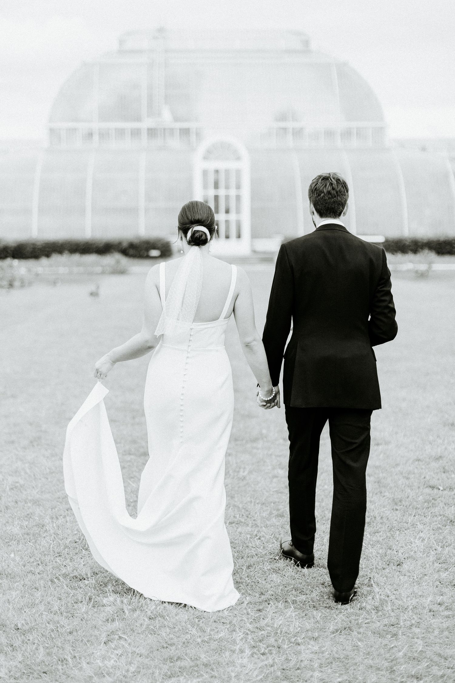A wedding couple walks towards a conservatory building in black and white photography.