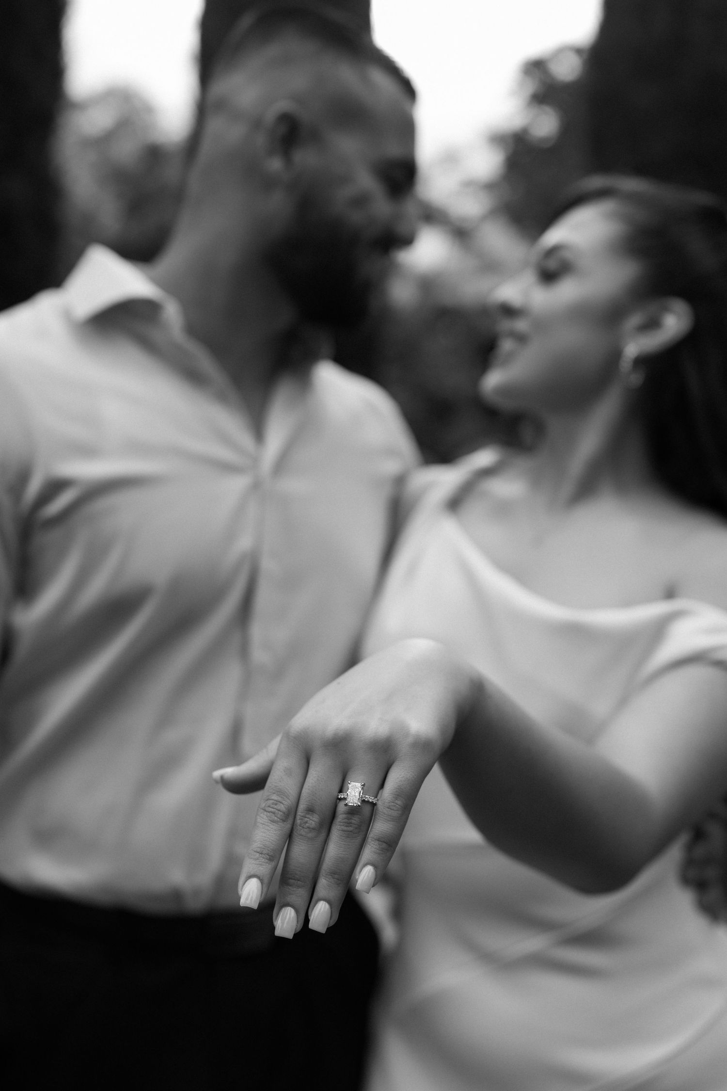 Black and white portrait of a couple showing off an engagement ring in a garden setting.