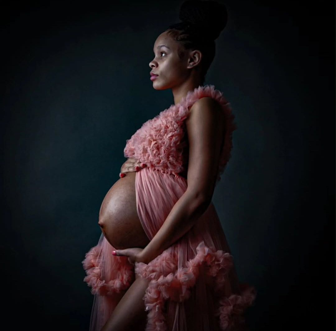 Pregnant woman in a pink ruffled tulle gown posing in profile against a dark moody background in an elegant maternity portrait.
