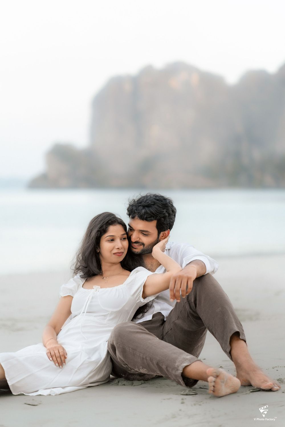 Couple relaxing on beach with cliffs in background during casual engagement photoshoot.