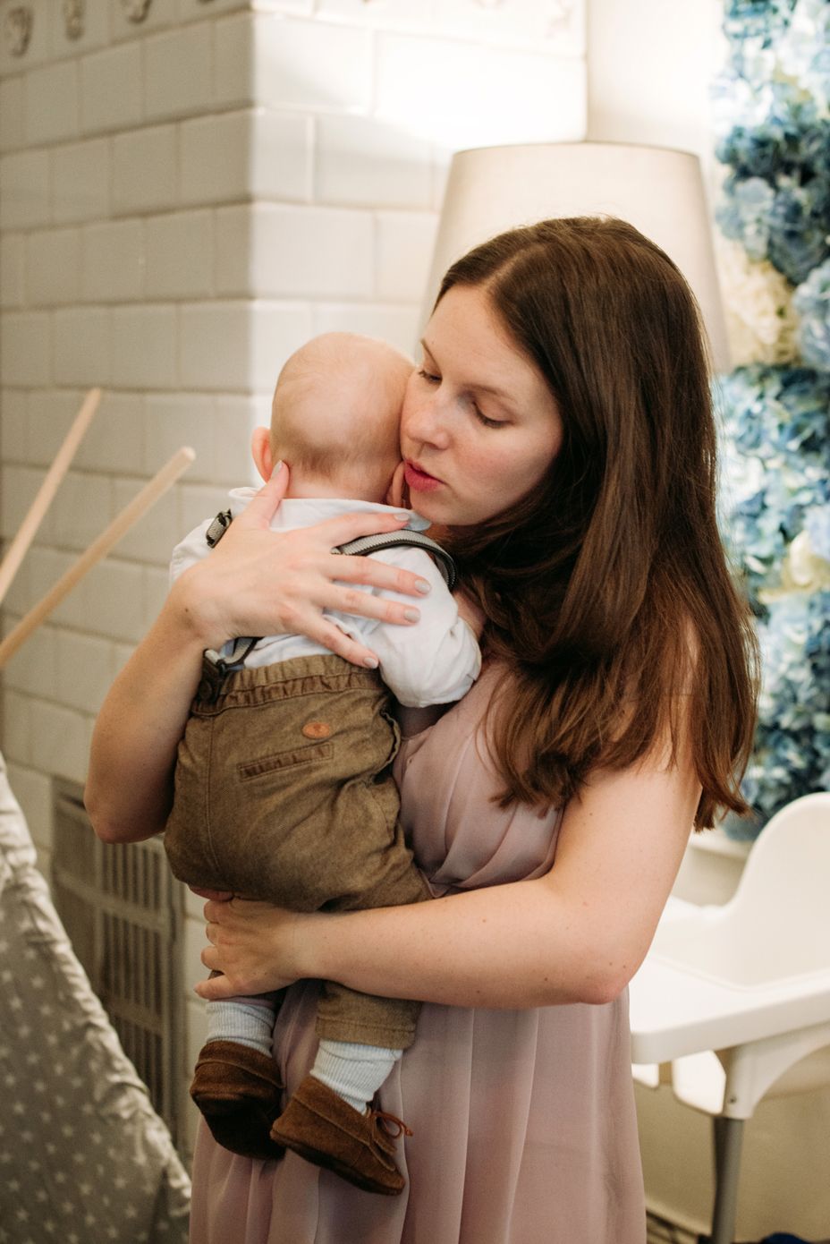 A woman in a pink dress holds and kisses a baby wearing brown overalls in a cozy home setting.