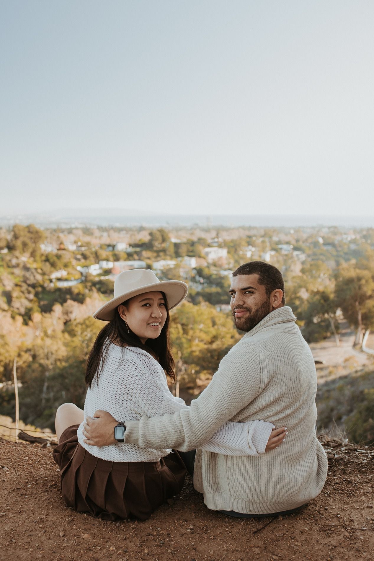 A couple in cozy white sweaters sits together on a hilltop overlooking the Los Angeles cityscape at sunset at Will Rogers State Park.