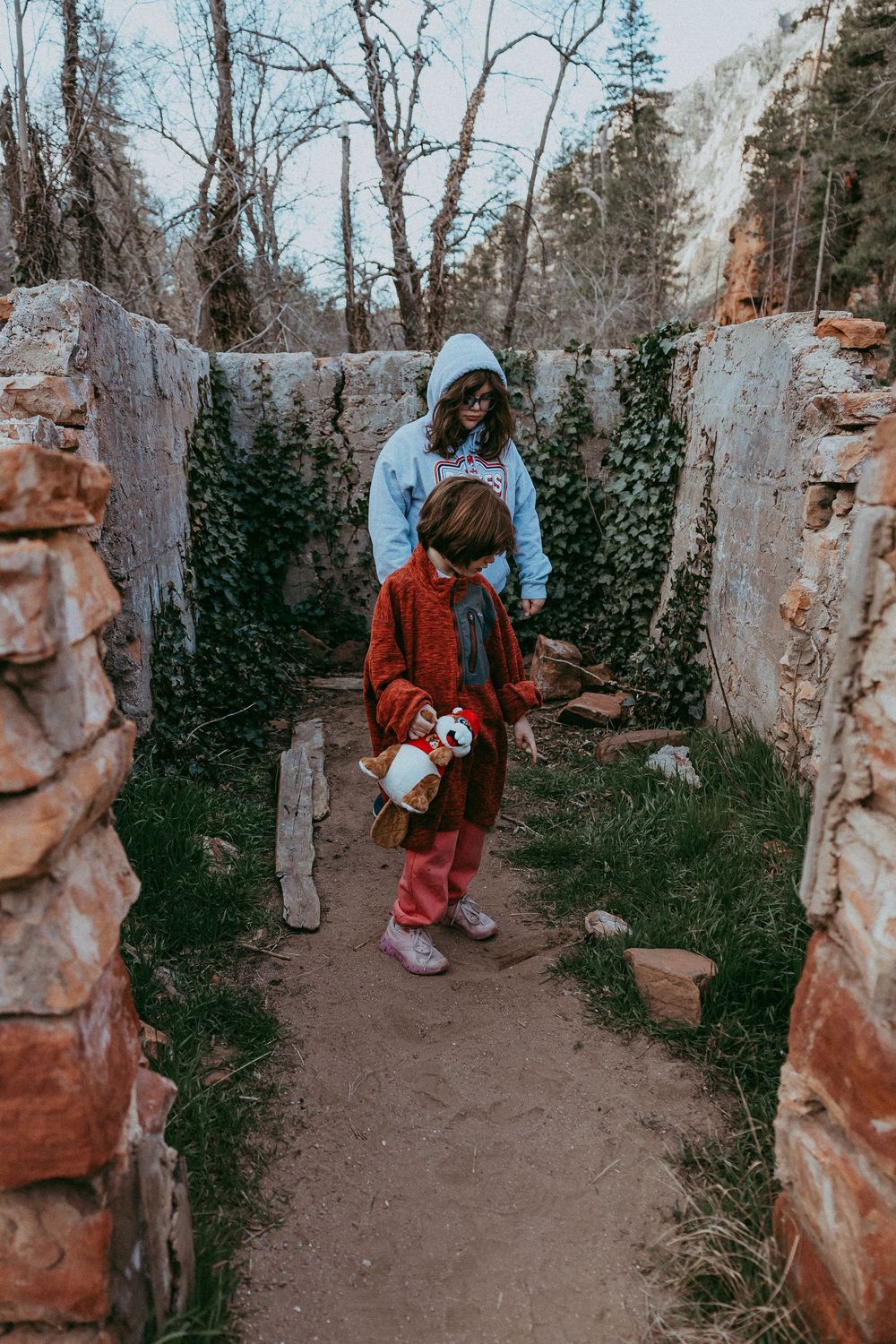Two children explore ancient stone ruins surrounded by ivy-covered walls and trees in a rustic outdoor setting.