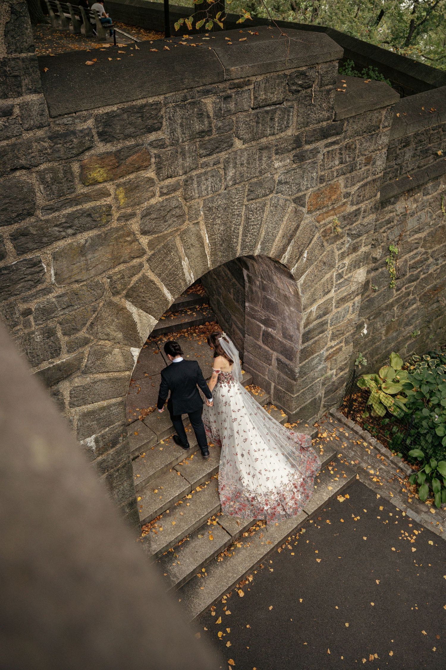 Bride and groom walking through stone archway with fallen autumn leaves.