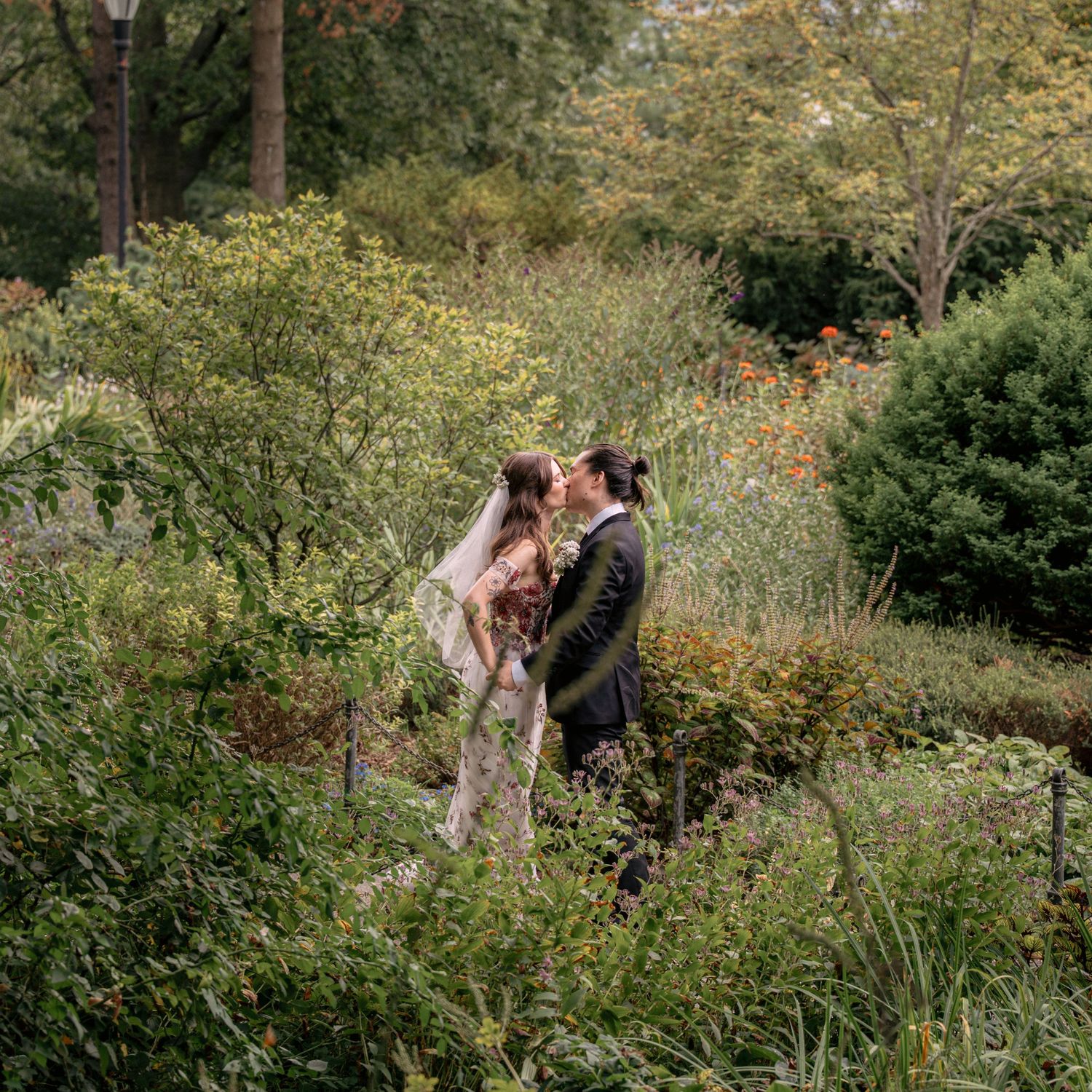 Bride and groom kissing among lush garden greenery during outdoor wedding.