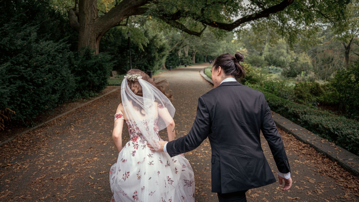Bride and groom walking hand in hand down a tree-lined garden path.