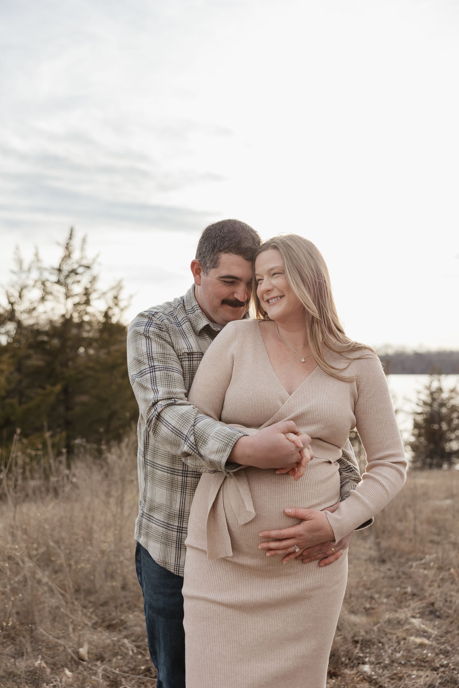 pregnant mother and dad to be holds baby bump in front of lake olathe during photo session as date night ideas in kansas city