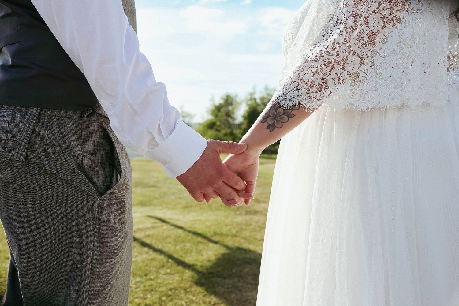 Close-up of bride and groom holding hands outdoors, showcasing lace wedding dress and tattooed arm in sunlight.