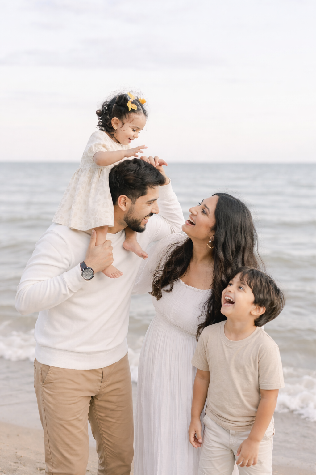 Happy mixed family of four laughing on beach with father lifting daughter on shoulders near ocean.