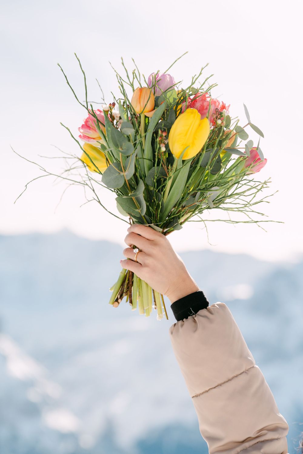 Hand in beige sweater sleeve holds up colorful bouquet of fresh flowers against bright blue sky background.