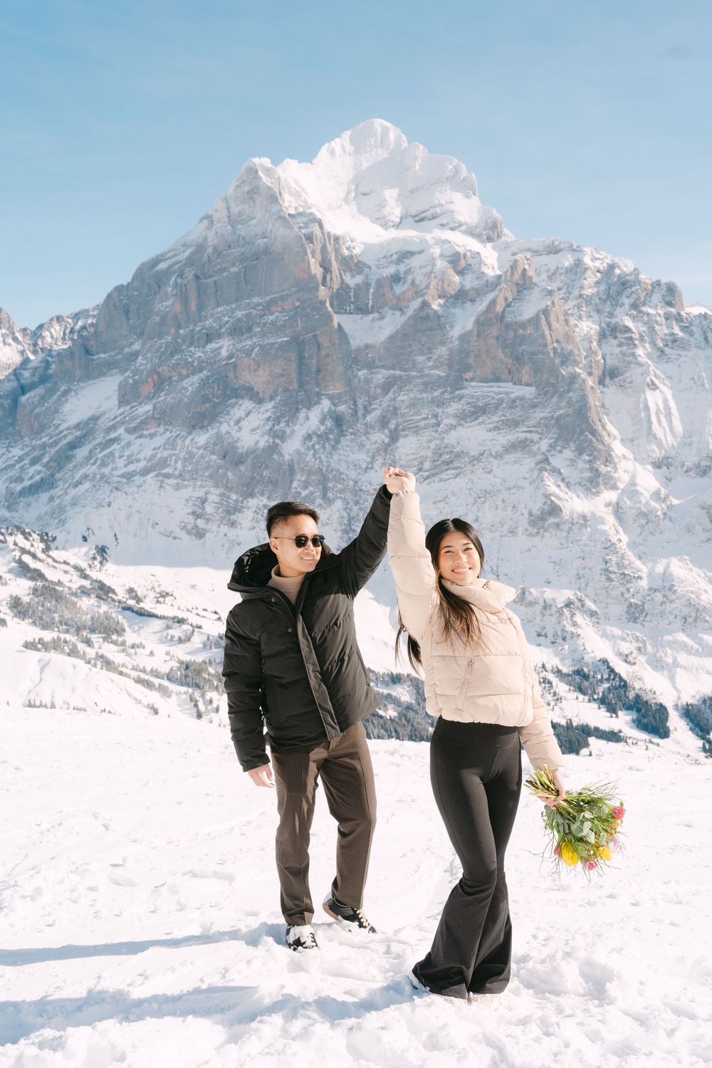 Two people playfully pose in the snow with dramatic snow-capped mountain peaks in the background.